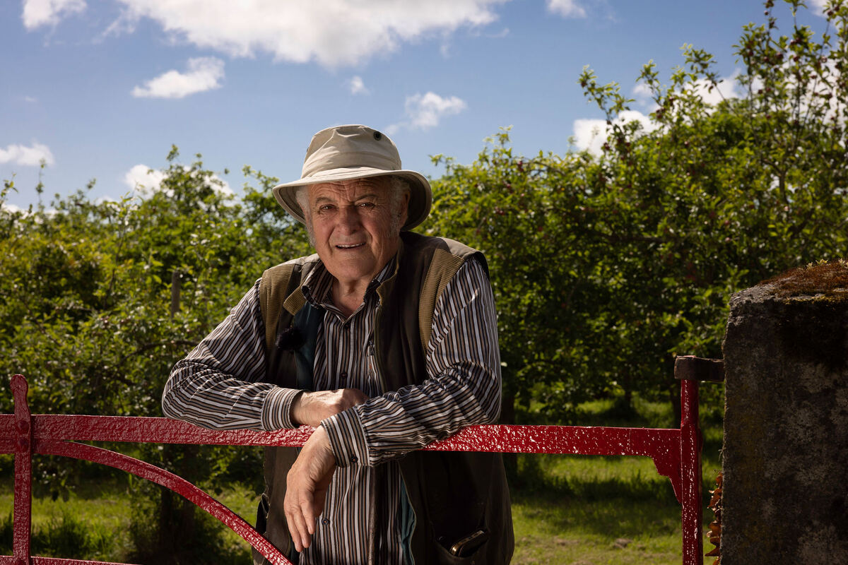 Rod from Highbank Orchards in hat and vest leaning on red gate in Orchard setting